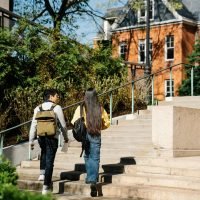Two young adults walking up outdoor stairs surrounded by greenery and buildings.